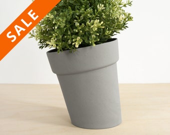 May include: A gray ceramic planter with a green leafy plant inside. The planter is tilted to the side.