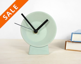 May include: A light green ceramic clock with black hands and a white face. The clock is on a light wood surface.