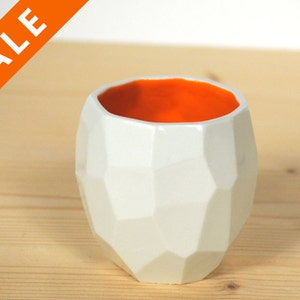 May include: White ceramic planter with a geometric design and an orange interior. The planter is sitting on a wooden surface.