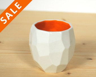 May include: White ceramic planter with a geometric design and an orange interior. The planter is sitting on a wooden surface.