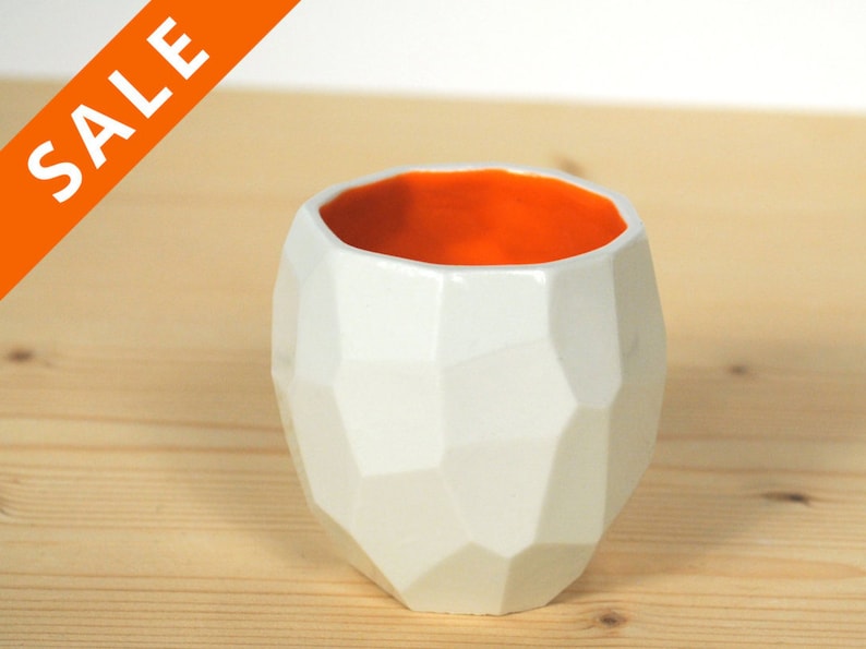 May include: White ceramic planter with a geometric design and an orange interior. The planter is sitting on a wooden surface.