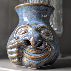 May include: A small, blue-glazed ceramic vessel featuring a sculpted face with a prominent nose, wide eyes, and a hand resting near the chin. The glaze has a mottled texture with darker brown accents highlighting the facial features and contours.