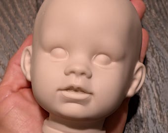 May include: A blank, unpainted doll head with closed eyes, a small nose, and a slightly open mouth. The doll head is held in a hand, showcasing its smooth, pale surface. The head is a light beige color and has a neck base.