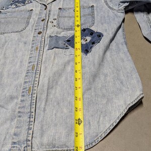May include: A light blue denim shirt with a patchwork design and silver studs. The shirt has a button-up front and long sleeves. The shirt is measured with a tape measure, showing a length of 68.58 cm.