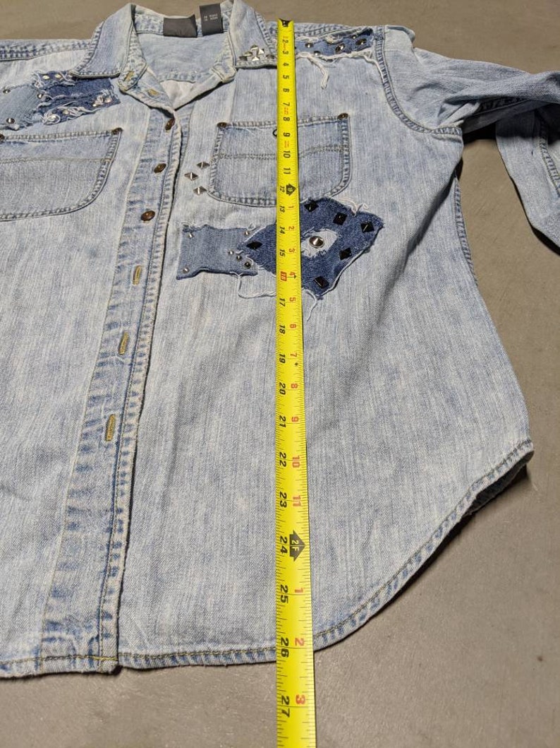May include: A light blue denim shirt with a patchwork design and silver studs. The shirt has a button-up front and long sleeves. The shirt is measured with a tape measure, showing a length of 68.58 cm.