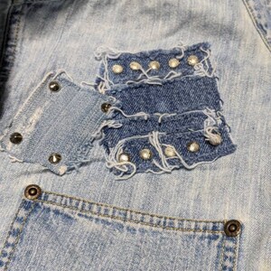 May include: Light blue denim jacket with a patch pocket on the front. The pocket is made of a different shade of denim and has silver studs around the edges.
