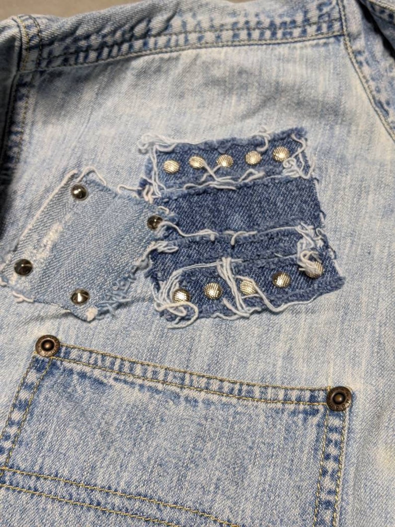 May include: Light blue denim jacket with a patch pocket on the front. The pocket is made of a different shade of denim and has silver studs around the edges.
