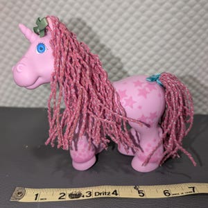 May include: A pink unicorn toy with a long, textured pink mane and tail, featuring blue eyes and a star pattern on its body. A green bow is on its head and a blue bow is on its tail. A yellow measuring tape shows the toy is approximately 7 inches long.