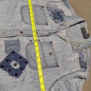 May include: Light blue denim jacket with studded details and patches. The jacket has a peace sign on the pocket and a measuring tape is used to show the size.