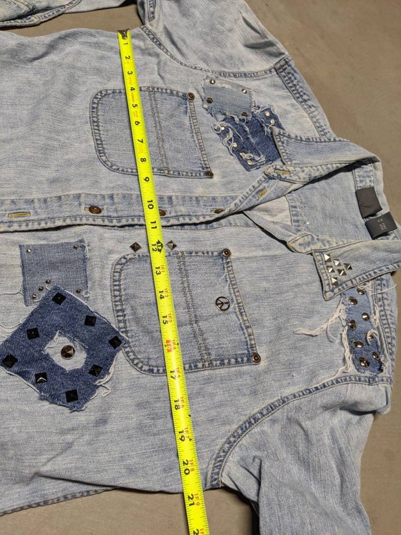 May include: Light blue denim jacket with studded details and patches. The jacket has a peace sign on the pocket and a measuring tape is used to show the size.