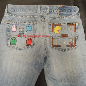 May include: Light blue denim jeans with a Pac-Man and Ms. Pac-Man design on the back pockets. The Pac-Man design includes a yellow Pac-Man, a brown maze, and red cherries. The Ms. Pac-Man design includes four ghosts in different colors: pink, blue, red, and green.