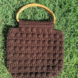 May include: A brown crocheted tote bag with a wooden handle. The bag has a textured, puffy design.
