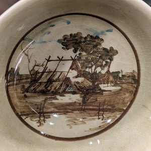 May include: A cream-colored ceramic bowl with a hand-painted landscape scene inside. The scene features a tree, water, and structures in shades of brown and tan, with blue accents. The bowl has a slightly cracked glaze.