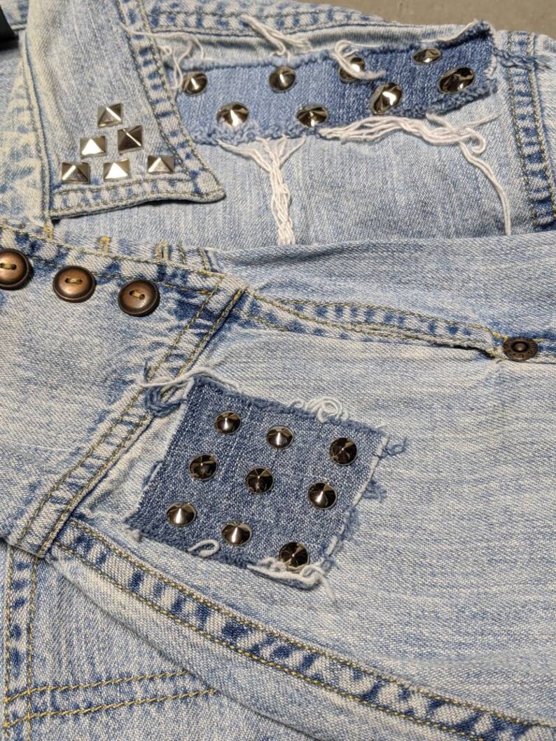 May include: Light blue denim jacket with multiple patches and silver studs. The patches are made of denim and have frayed edges. The jacket has a collar and a button closure.