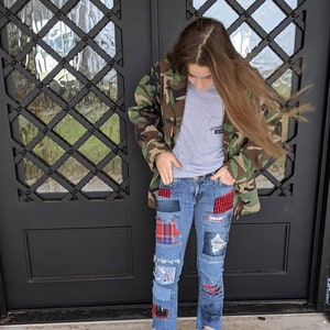 May include: A person wearing a camouflage jacket, a grey t-shirt with the text "Good Vibes 18-76", and blue jeans with patches. The jeans have red and black plaid patches, and the person is wearing black ankle boots.