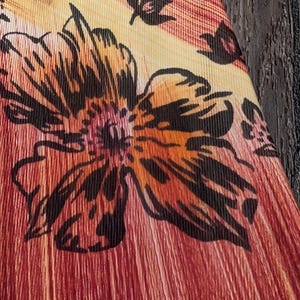 May include: Close-up of a fabric with a floral pattern. The design features a large, stylized flower in shades of orange, brown, and black, set against a background of red and yellow stripes. The fabric has a textured, ribbed appearance.