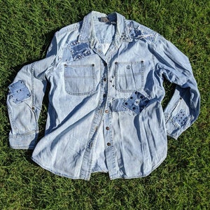 May include: A light blue denim shirt with patches and studs. The shirt has a button-down front and long sleeves.