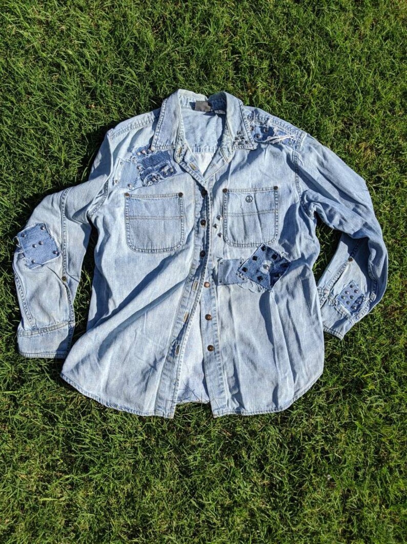 May include: A light blue denim shirt with patches and studs. The shirt has a button-down front and long sleeves.