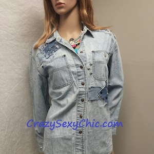 May include: Light blue denim shirt with distressed details, including patches and studs. The shirt has a button-down front and long sleeves. The text "CrazySexyChic.com" is visible on the bottom of the shirt.