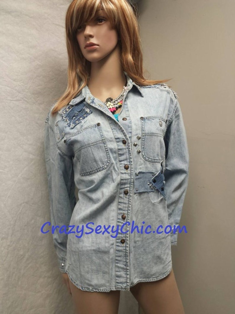 May include: Light blue denim shirt with distressed details, including patches and studs. The shirt has a button-down front and long sleeves. The text "CrazySexyChic.com" is visible on the bottom of the shirt.