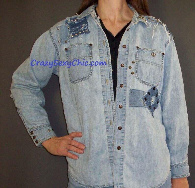 May include: Light blue denim shirt with a patchwork design and silver studs. The shirt has a button-up front and long sleeves. The shirt is distressed and has a vintage look.