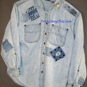 May include: A light blue denim shirt with multiple patches and studs. The shirt has a button-down front and long sleeves. The text "CrazySexyChic.com" is printed on the front of the shirt.