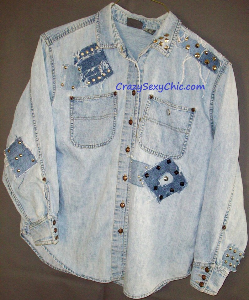 May include: A light blue denim shirt with multiple patches and studs. The shirt has a button-down front and long sleeves. The text "CrazySexyChic.com" is printed on the front of the shirt.