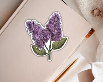 Könnte beinhalten: Ein gestanzter Aufkleber mit einer Aquarell-Illustration von zwei violetten Fliederblüten-Clustern mit grünen Blättern und einer dunklen Umrandung. Der Aufkleber liegt auf einem hellrosa Notizbuch neben einem goldenen Stift und einem goldenen Ring.