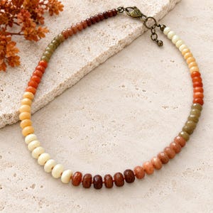 May include: A beaded necklace featuring a gradient of smooth, round gemstone beads in shades of cream, yellow, orange, red, and brown. The necklace is secured with a bronze-toned metal lobster clasp and an adjustable chain with a small heart charm.
