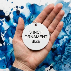 Puede incluir: Un adorno circular de cer&aacute;mica blanca con el texto negro 3 INCH ORNAMENT SIZE impreso en el centro. El adorno incluye una peque&ntilde;a anilla de metal plateado en la parte superior para colgarlo, proporcionando una referencia visual clara de las medidas.