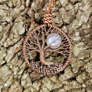 May include: A copper-colored wire-wrapped pendant featuring a tree of life design. The pendant has a circular frame with a detailed tree silhouette and a light blue gemstone. The pendant hangs on a matching copper chain.