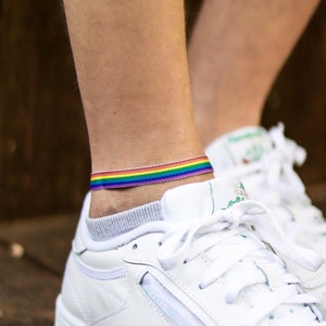 May include: White leather sneakers with a rainbow ankle bracelet. The sneakers have white laces and a green logo on the side.