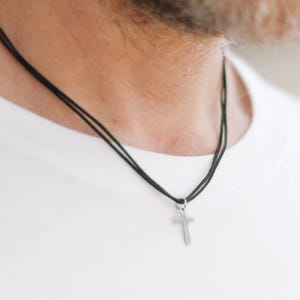 May include: A silver cross pendant hangs from a black cord necklace. The necklace is worn over a white t-shirt. The image is a close-up of the necklace and upper chest.