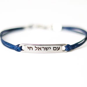 May include: A dark blue cord bracelet featuring a central silver-toned metal bar engraved with the Hebrew text "עם ישראל חי". The cord is thin and circular, connecting to the metal plate on both sides with a simple knot design.