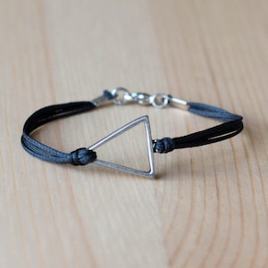 Puede incluir: Una pulsera minimalista con un colgante de triángulo plateado. La pulsera tiene un diseño de cordón negro y gris con un cierre plateado. La joya se exhibe sobre una superficie de madera clara.