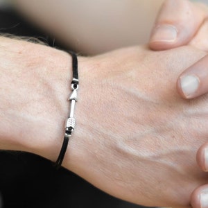 May include: A black cord bracelet with a silver-colored arrow-shaped charm is worn on a person's wrist. The bracelet is the focal point, set against a neutral background. The person's hands are clasped together.