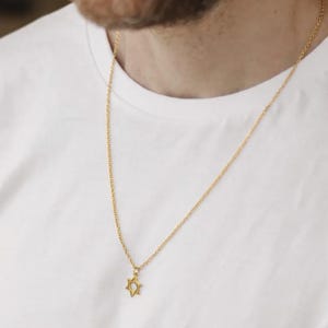 May include: A gold-colored necklace with a Star of David pendant hangs against a white t-shirt. The chain is delicate, and the pendant is a six-pointed star. The necklace is a religious or fashion accessory.