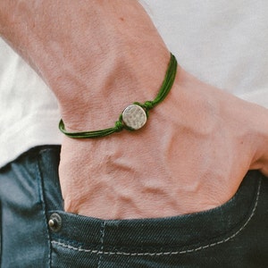 Könnte beinhalten: Ein Armband bestehend aus einer dünnen, dunkelgrünen Kordel aus mehreren Strängen und einem einzelnen, kleinen, kreisförmigen, silberfarbenen Metallanhänger mit einer texturierten, gehämmerten Oberfläche.