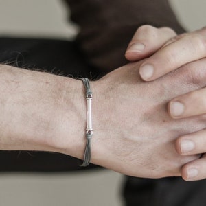 Puede incluir: Una pulsera minimalista con una cuenta cilíndrica central de color claro y detalles plateados. La pulsera está ensartada en múltiples hebras de cordón gris oscuro. La imagen muestra la pulsera en la muñeca de una persona.