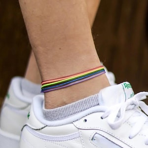 Puede incluir: Zapatillas blancas de cuero con cordones blancos y un logotipo de Reebok verde. Las zapatillas son usadas por una persona que lleva un brazalete de tobillo de colores arcoíris.