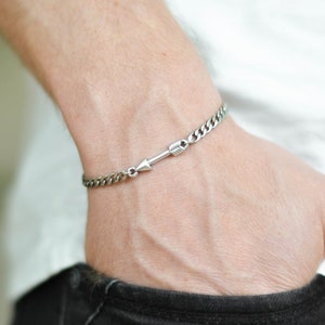 May include: A silver chain bracelet with an arrow charm. The bracelet is on a person's wrist.