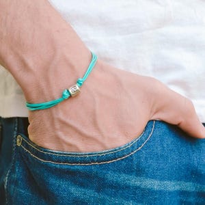May include: A turquoise blue string bracelet with a silver charm featuring the Cancer zodiac symbol.