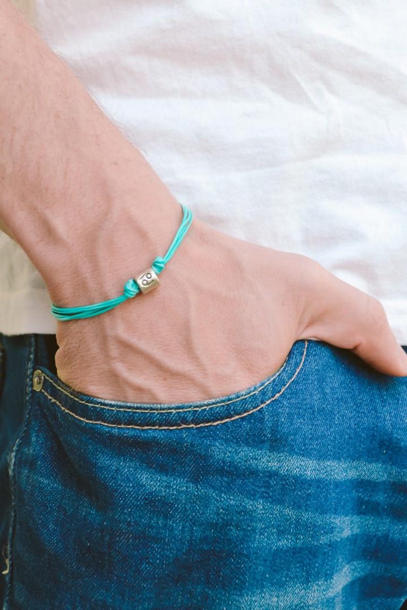 May include: A turquoise blue string bracelet with a silver charm featuring the Cancer zodiac symbol.