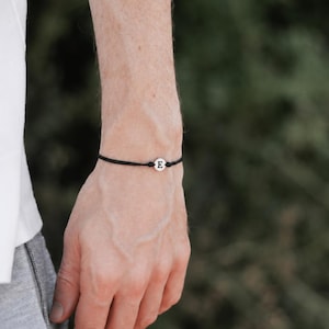 Könnte beinhalten: Ein schwarzes Kordelarmband mit einem kleinen, runden silberfarbenen Anhänger, auf dem der Großbuchstabe E in Schwarz eingraviert ist. Das Armband ist dünn und minimalistisch und mit einer verstellbaren Kordelkonstruktion versehen.