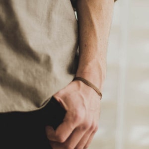 Op de afbeelding: Een persoon draagt een lichtbruin shirt met een bruin leren armband om de pols.