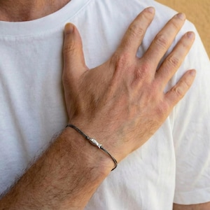 May include: A dark grey bracelet with a silver-coloured cylindrical bead is worn on a wrist. The bracelet is made of a thin, woven material. The person is wearing a white t-shirt.