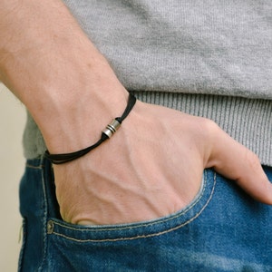 May include: Black bracelet with a silver cylindrical bead. The bracelet is worn on a wrist, with the person wearing a gray sweater and blue jeans. The bracelet is a simple, minimalist design.