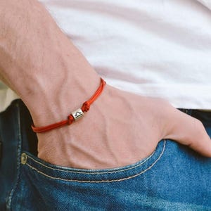 May include: A red string bracelet with a silver charm featuring the Libra zodiac symbol.