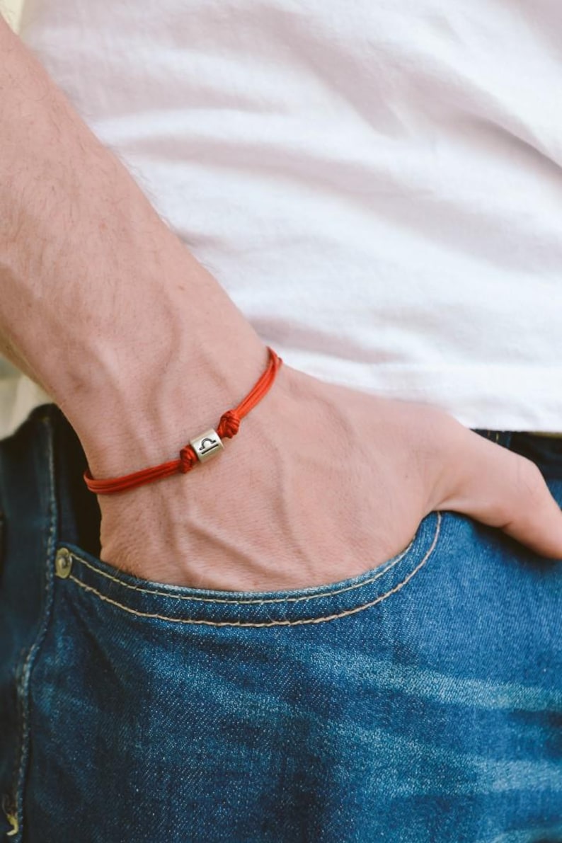 May include: A red string bracelet with a silver charm featuring the Libra zodiac symbol.