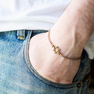 May include: A gold Star of David charm bracelet on a brown cord. The bracelet is worn on a person's wrist.